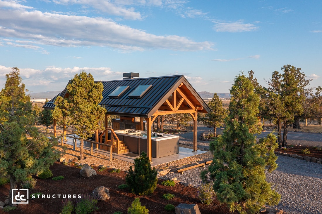 A small modern cabin with a metal roof and wooden beams sits among trees. A hot tub is on the deck, mountains in the background.