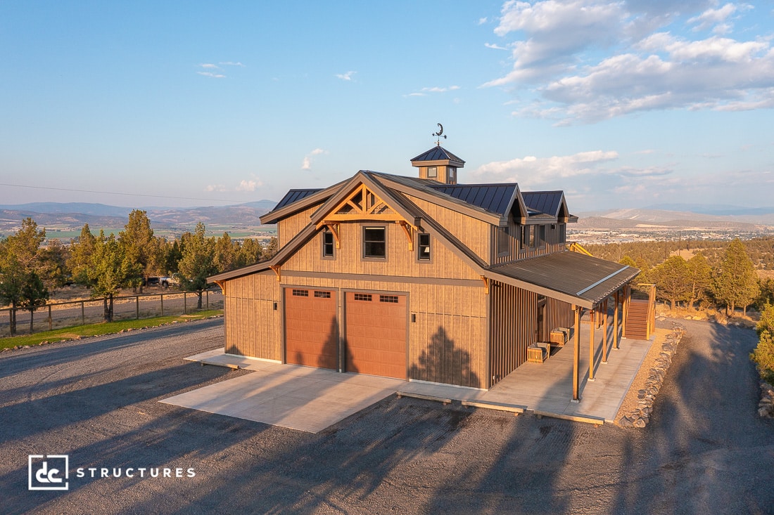 A large, modern barn-style building with brown siding, two red doors, and a cupola sits on gravel in a rural landscape.