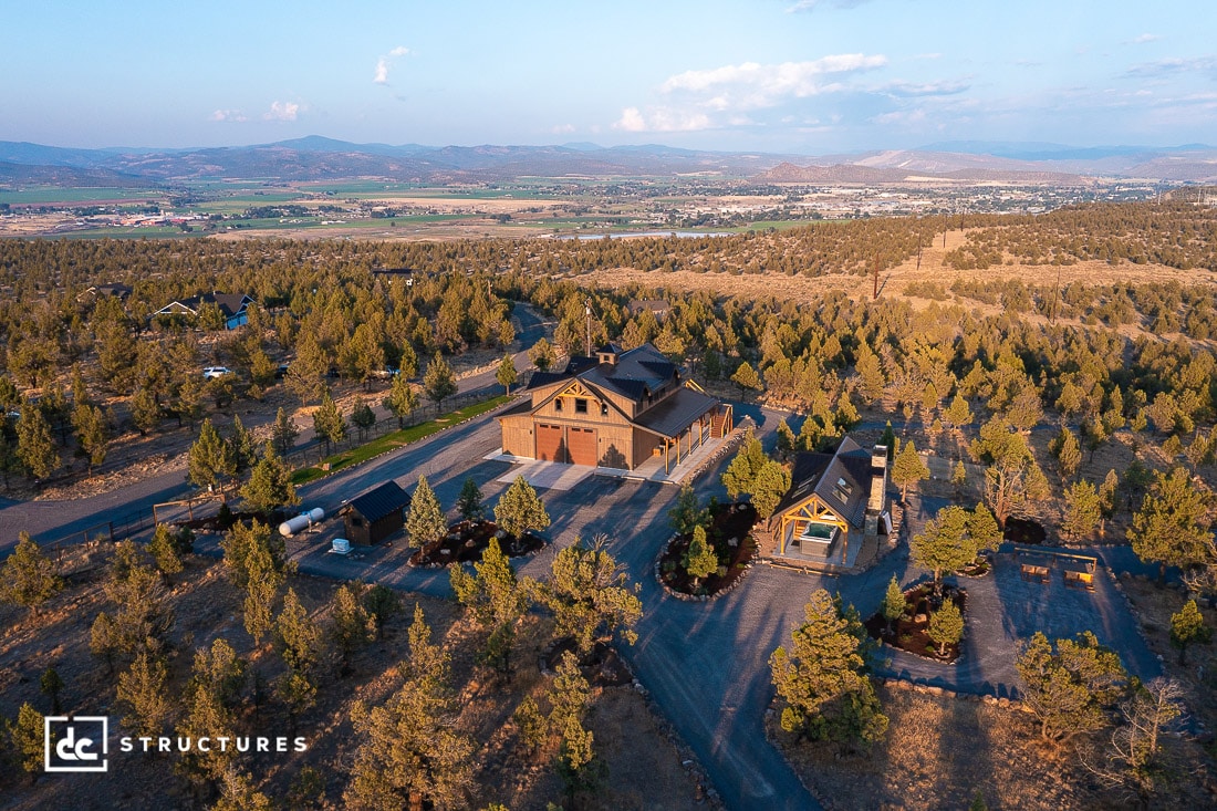 Aerial view of a large rustic barn-style house surrounded by trees with mountains and a town in the distance under a blue sky.