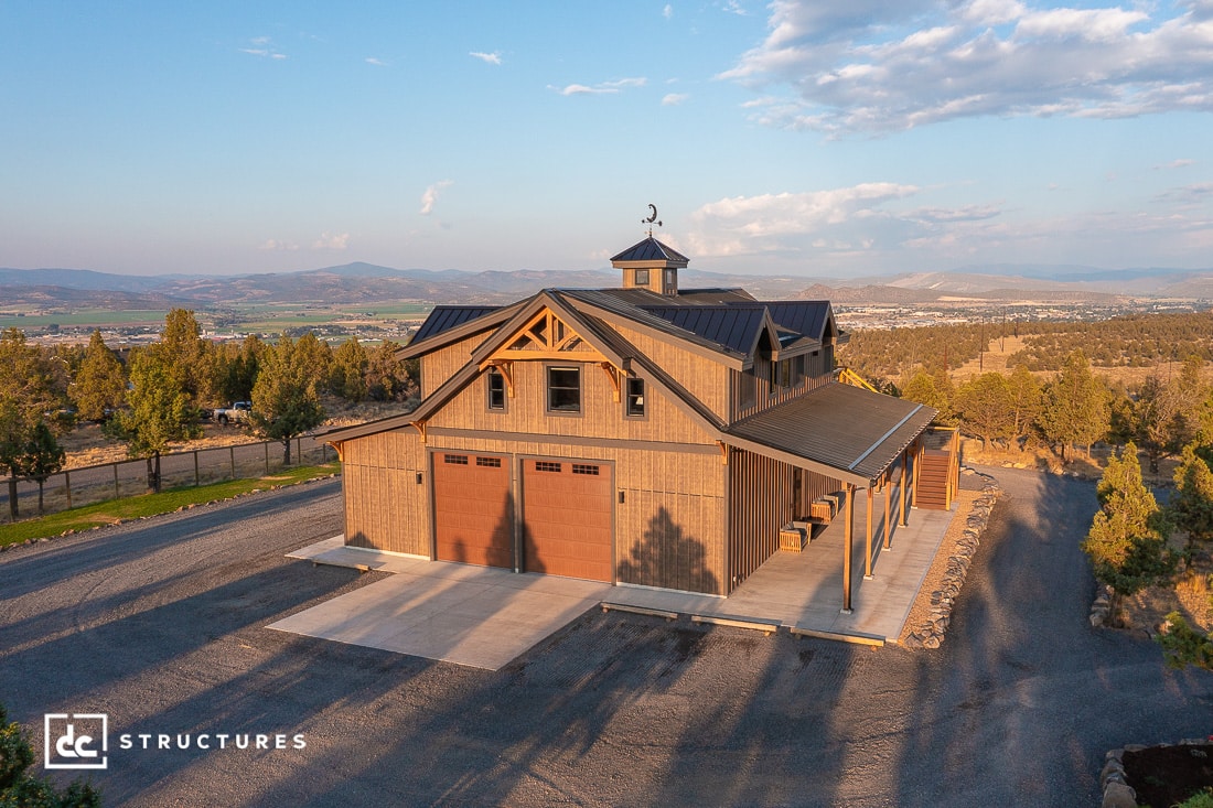 A large, modern two-story barn with three garage doors and a cupola sits in a rural landscape, surrounded by trees, hills, and open sky at sunset.