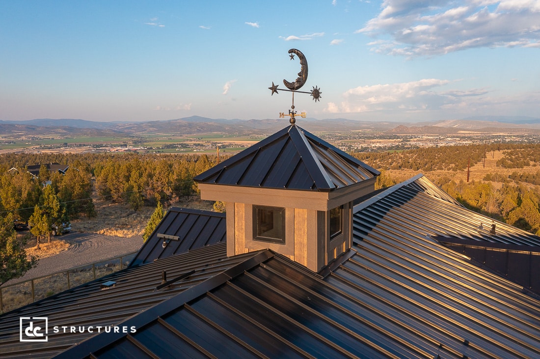 A metal-roofed building with a cupola and crescent moon and star weather vane overlooks hills, trees, and a distant valley.