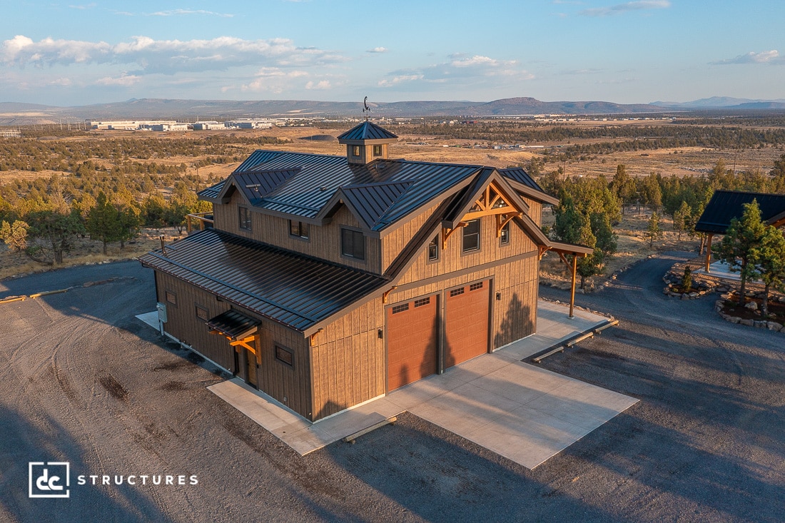 A large rustic barn-style building with three garage doors and a cupola in a rural landscape of trees, open land, and distant hills.