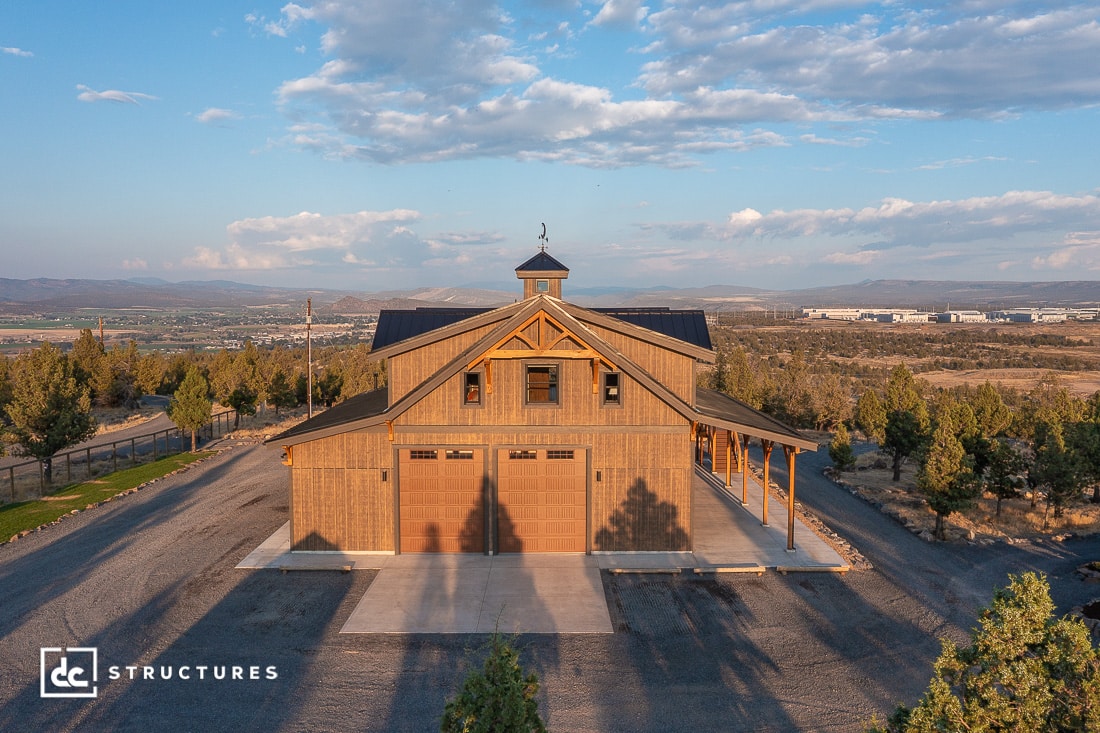 Aerial view of a large wooden barn with a cupola, surrounded by trees and mountains under a blue sky, with garage doors and porch.