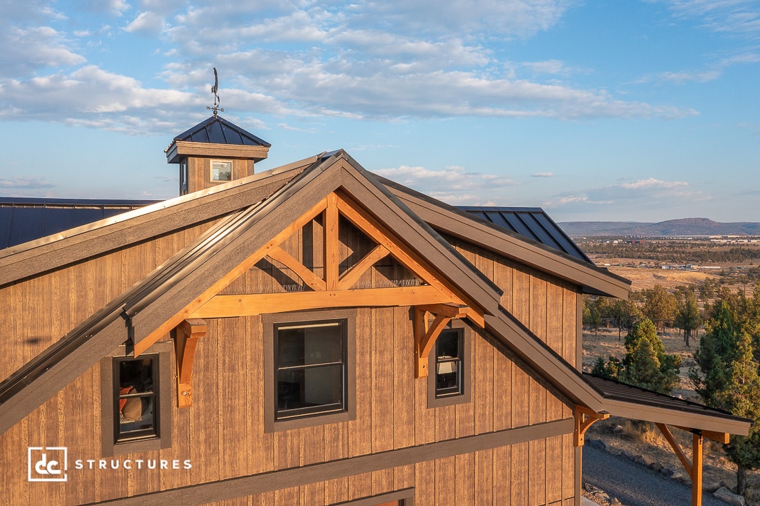 A wooden barn with a metal roof and large windows sits on a hillside, with a clear sky and wide landscape in the background.