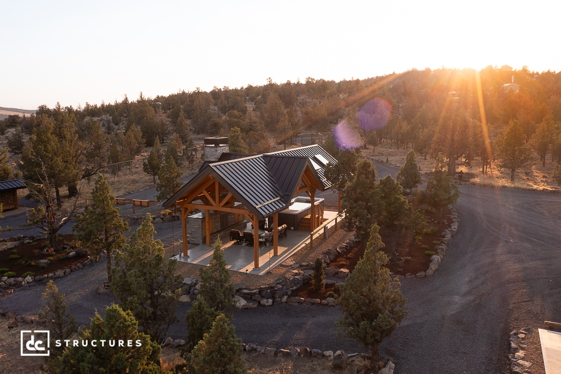A wooden pavilion with a black metal roof sits among trees and landscaped paths, illuminated by the golden light of the setting sun in a rural, rocky area.