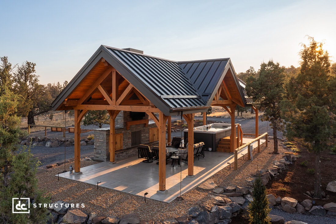 A wooden outdoor pavilion with a metal roof, open sides, concrete floor, hot tub, lounge chairs, and stone barbeque at sunset.