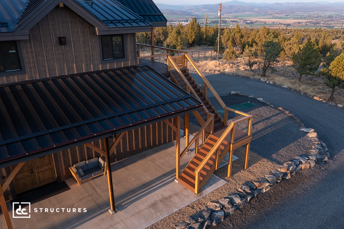 A modern two-story house with a metal roof, wood paneling, and a large wooden staircase leading to an upper deck in a rural setting.