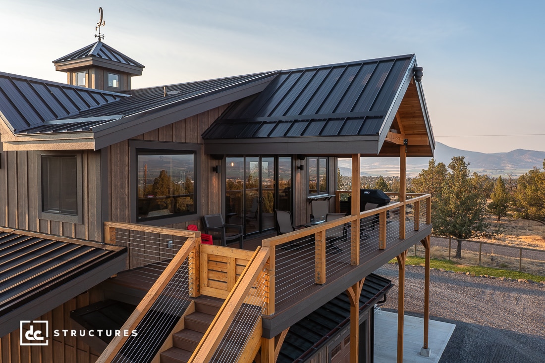 A modern wooden house with a large balcony and glass doors overlooks a mountainous landscape, with seating on the balcony.