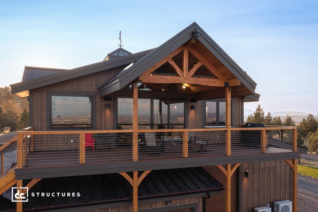 A modern two-story wooden cabin with large windows and a balcony, exposed beams, cable railing, set near trees at sunset.
