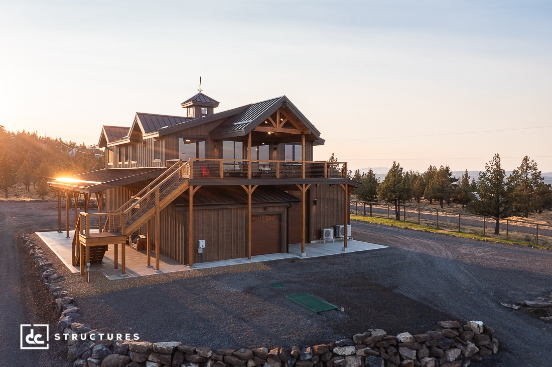 A two-story wooden barn-style house with large windows, a balcony, and an external staircase sits on a gravel lot in morning sunlight.