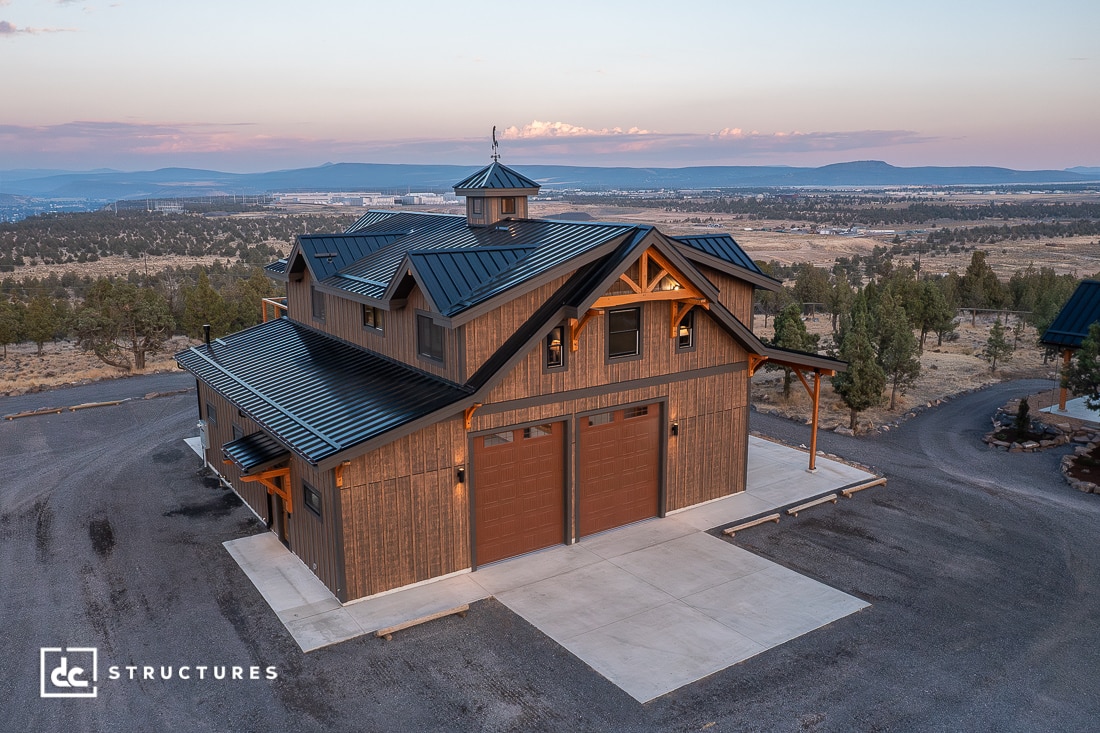 A large rustic barn-style building with a metal roof and wooden exterior sits on a concrete pad in a rural sunset landscape.