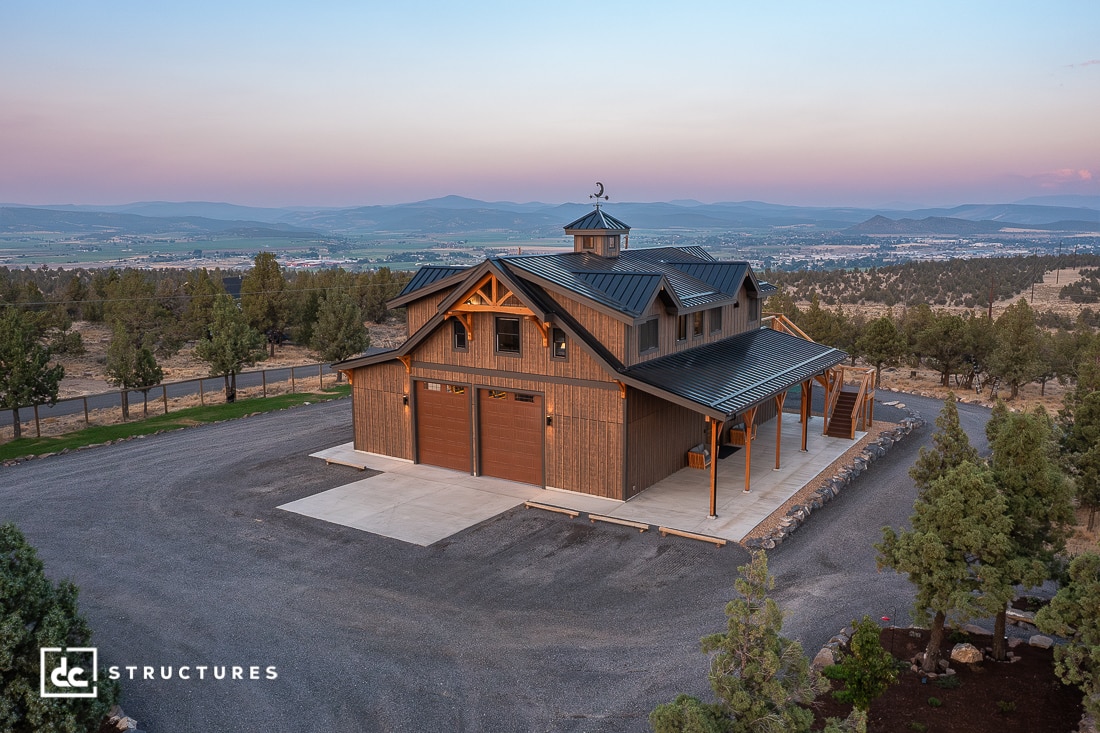 A large, rustic barn-style building with a metal roof and wooden exterior on a gravel lot, surrounded by trees at sunset.