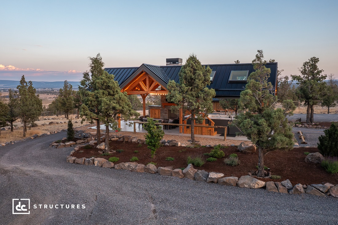 A modern barn-style home with a dark metal roof and timber accents sits among trees, landscaping, gravel paths, and countryside at sunset.