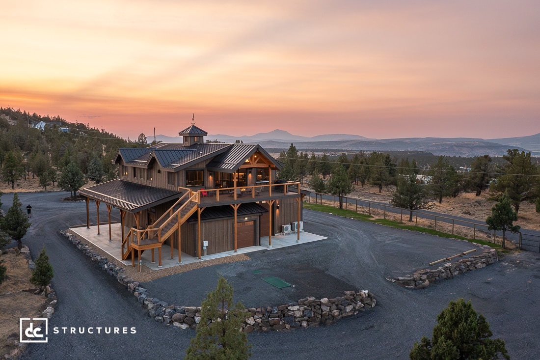 A large modern barn-style house with a wraparound deck sits on a spacious lot, trees, mountains, and a colorful sunset sky.