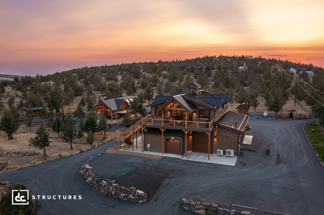 A modern two-story barn-style home with wood and metal accents sits among trees under a colorful sunset sky, surrounded by gravel driveways and outbuildings.