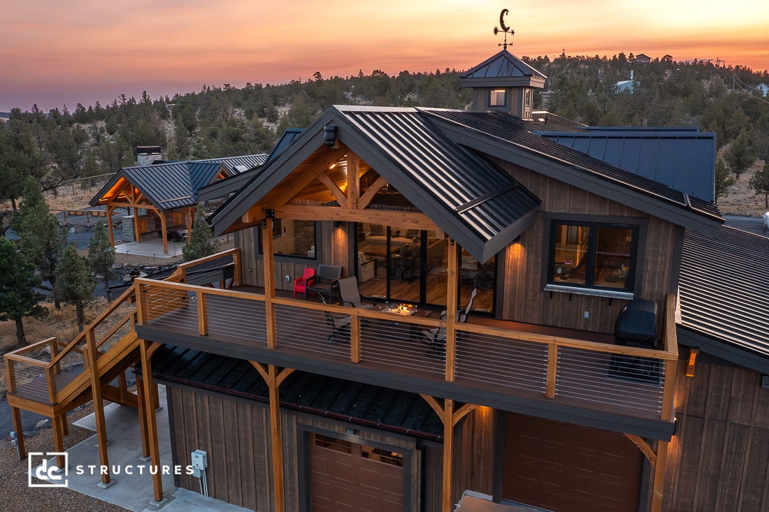 A modern two-story wooden house with large windows and a spacious balcony amid trees at sunset, metal roof, outdoor seating, warm lights.