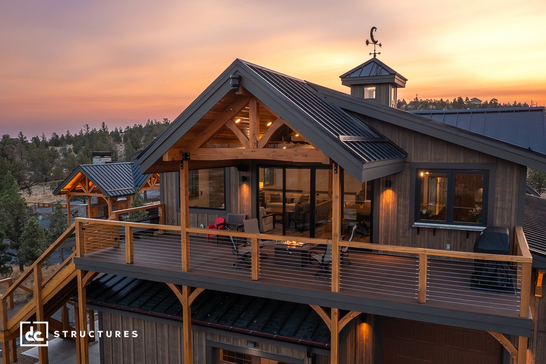 A modern wooden house with large windows, a spacious balcony, exposed beams, and cable railing against a sunset and forested hills.