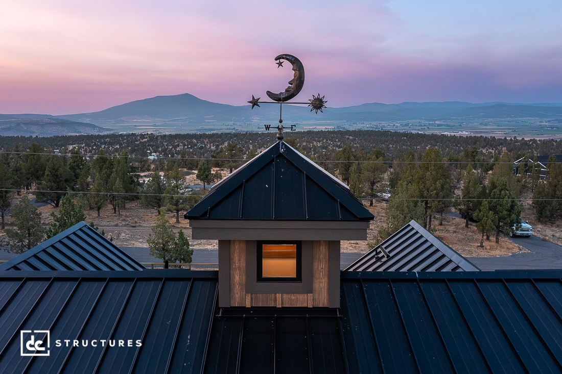 A rooftop cupola with a crescent moon and star weathervane overlooks a valley with trees and distant mountains at sunset. The sky is pink and purple.