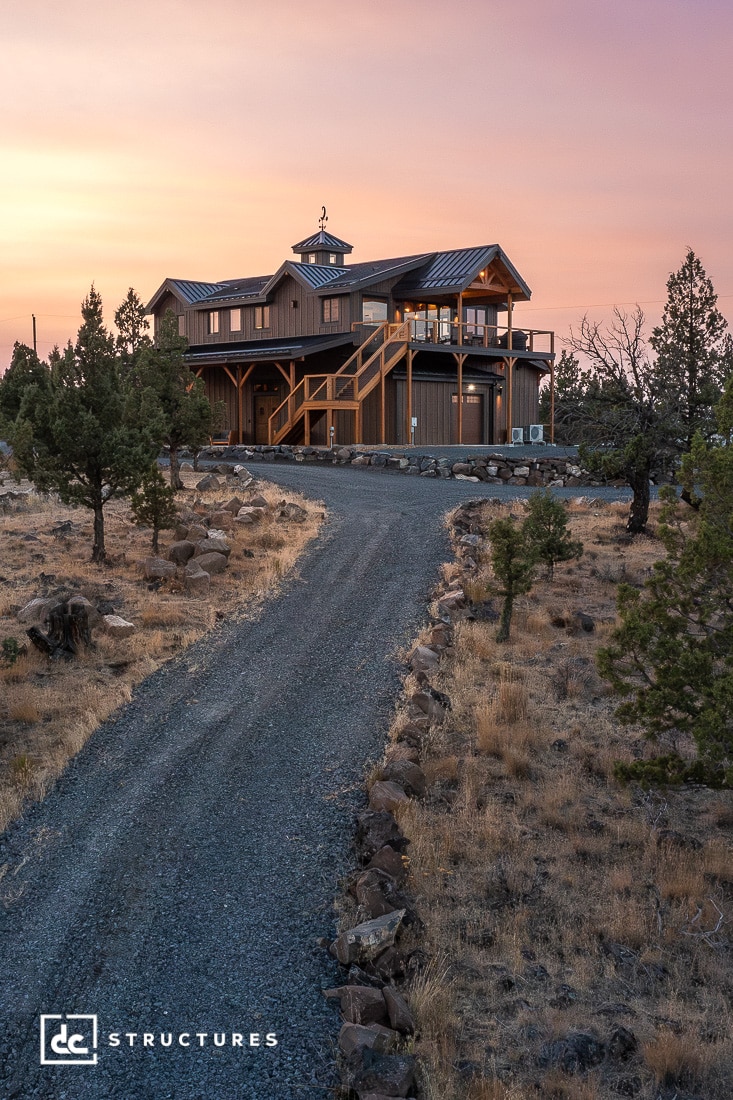 A modern wooden house with a wraparound porch sits on a rocky hill, surrounded by dry grass and trees under a sunset sky.