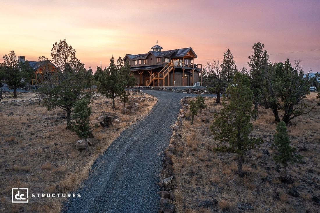 A gravel driveway winds through dry grass and scattered trees toward a two-story wooden house with a raised deck at sunset.