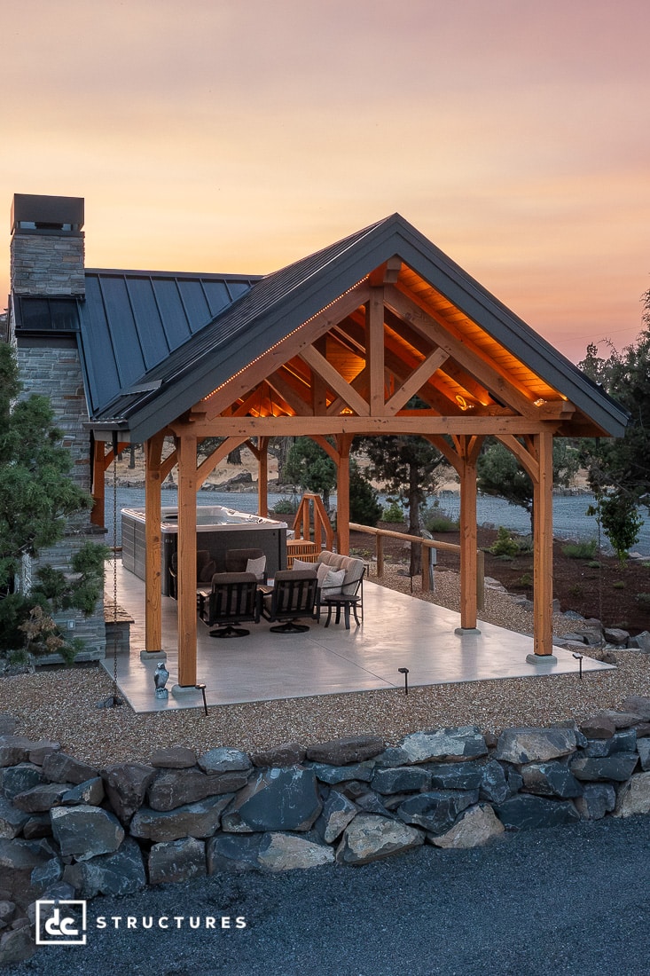A modern outdoor pavilion with wooden frame and metal roof, stone fireplace, dining table, lounge chairs, sunset sky, trees, stones.