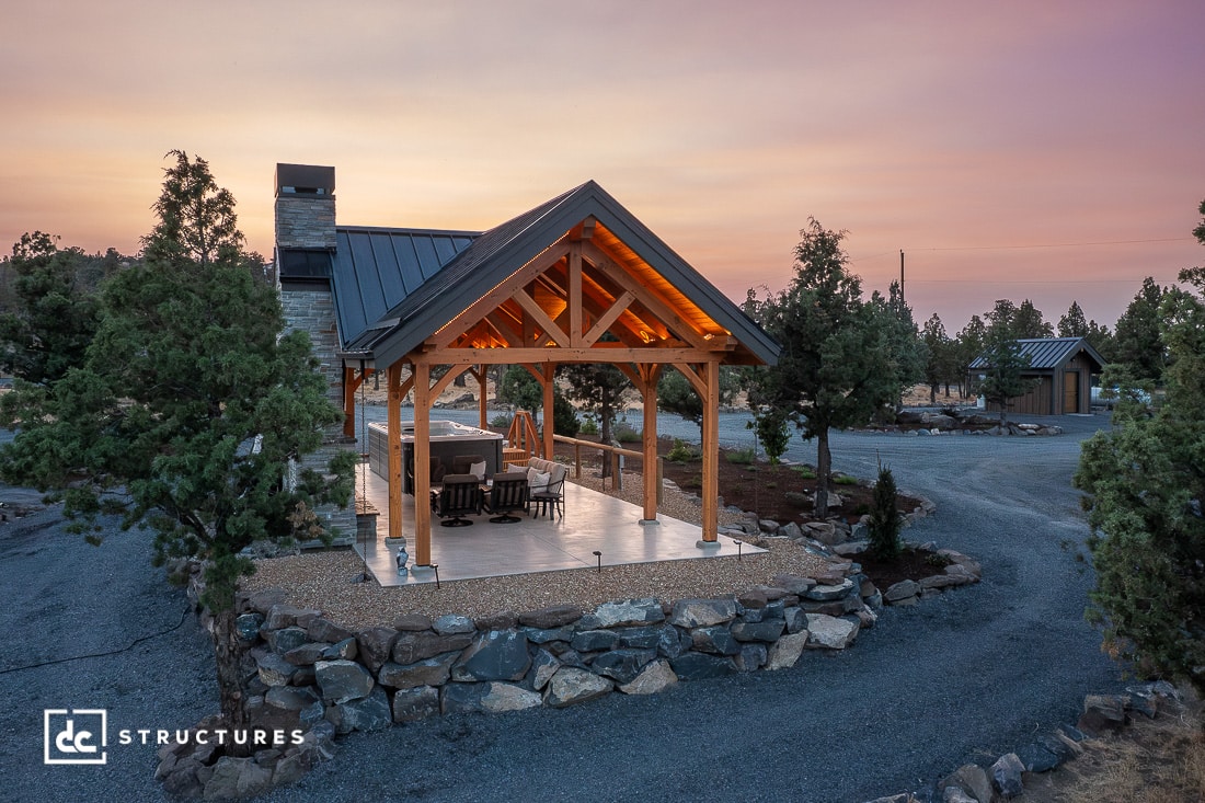 A cozy outdoor pavilion with a peaked wooden roof, glowing warmly at dusk amid trees and gravel paths. A small building is behind.