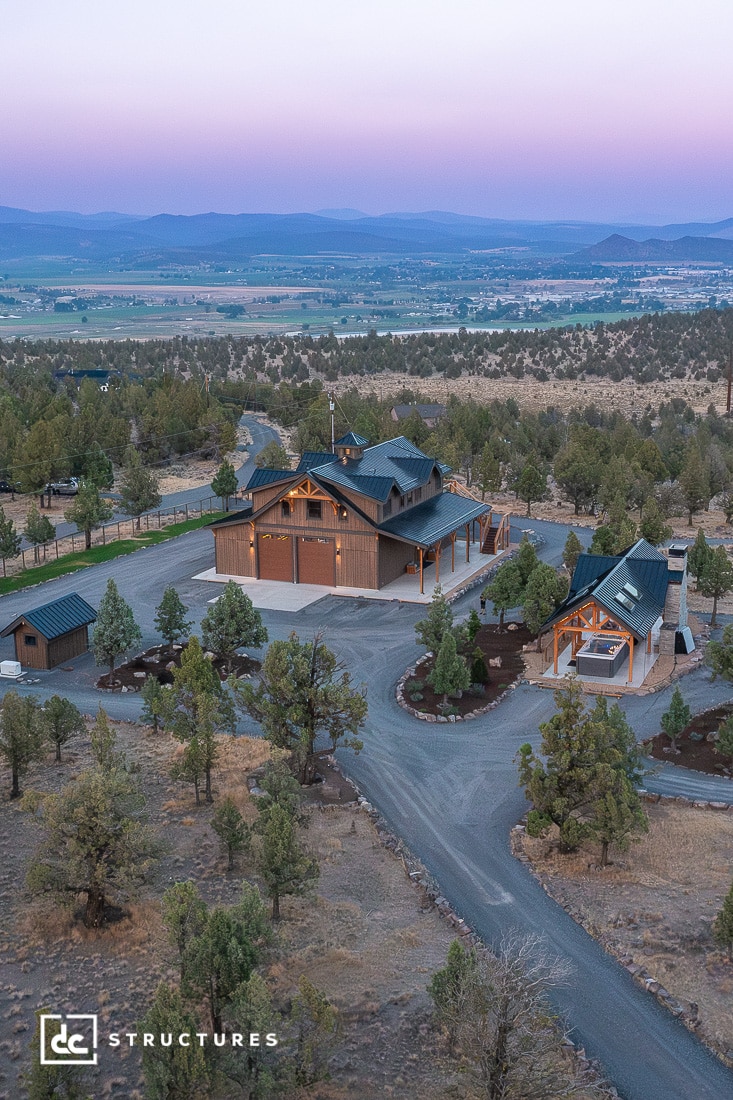 Aerial view of a large barn-style home with multiple peaked roofs, surrounded by trees, winding roads, and open fields at sunset.