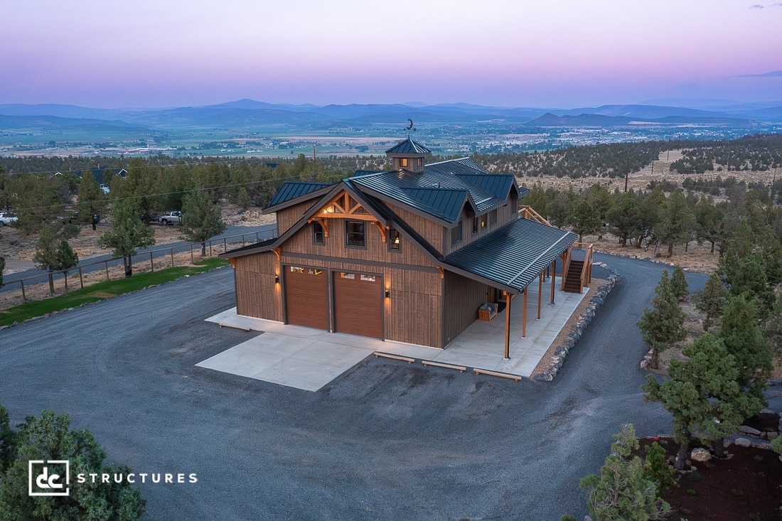 A large two-story barn-style building with green metal roofing and wood siding sits on a gravel lot surrounded by trees at sunset.
