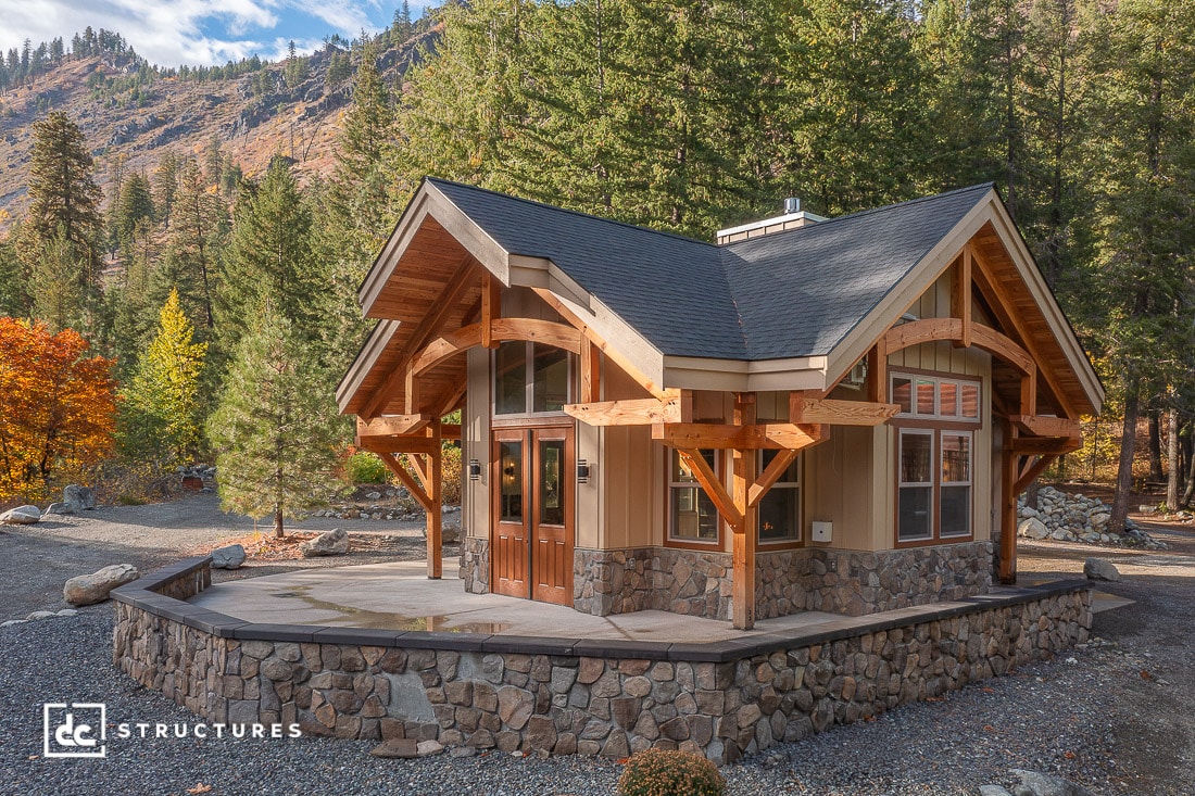 A small modern cabin with timber framing, stone foundation, and large windows sits among trees and mountains under a partly cloudy sky.