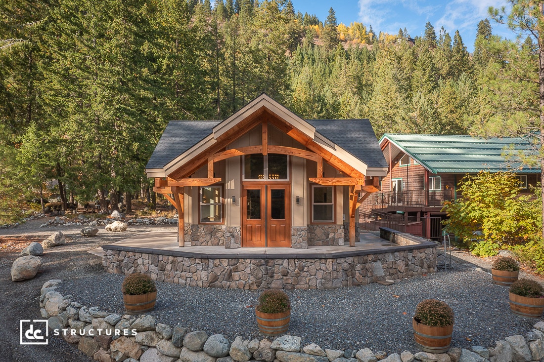 A small house with stone and wood exterior sits among trees and mountains. Potted plants line the patio, with another cabin nearby.