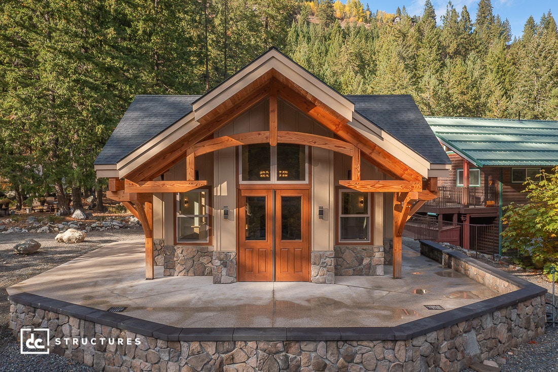 A small rustic cabin with stone and wood details, large front windows, and a peaked roof sits amid trees and a low stone wall.