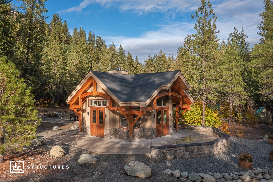 A small rustic cabin with a dark roof and wooden beams sits among tall pine trees and rocks under a partly cloudy sky. Peaceful scene.
