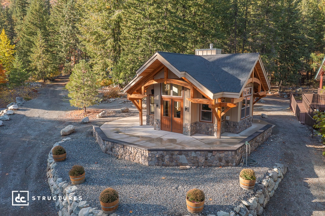 A small modern cabin with a stone foundation and wooden beams sits in a forest clearing with trees, gravel driveway, and potted plants.