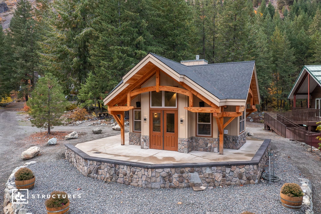 A small modern cabin with large windows, wooden beams, and a stone base sits among trees and gravel, with mountains behind.