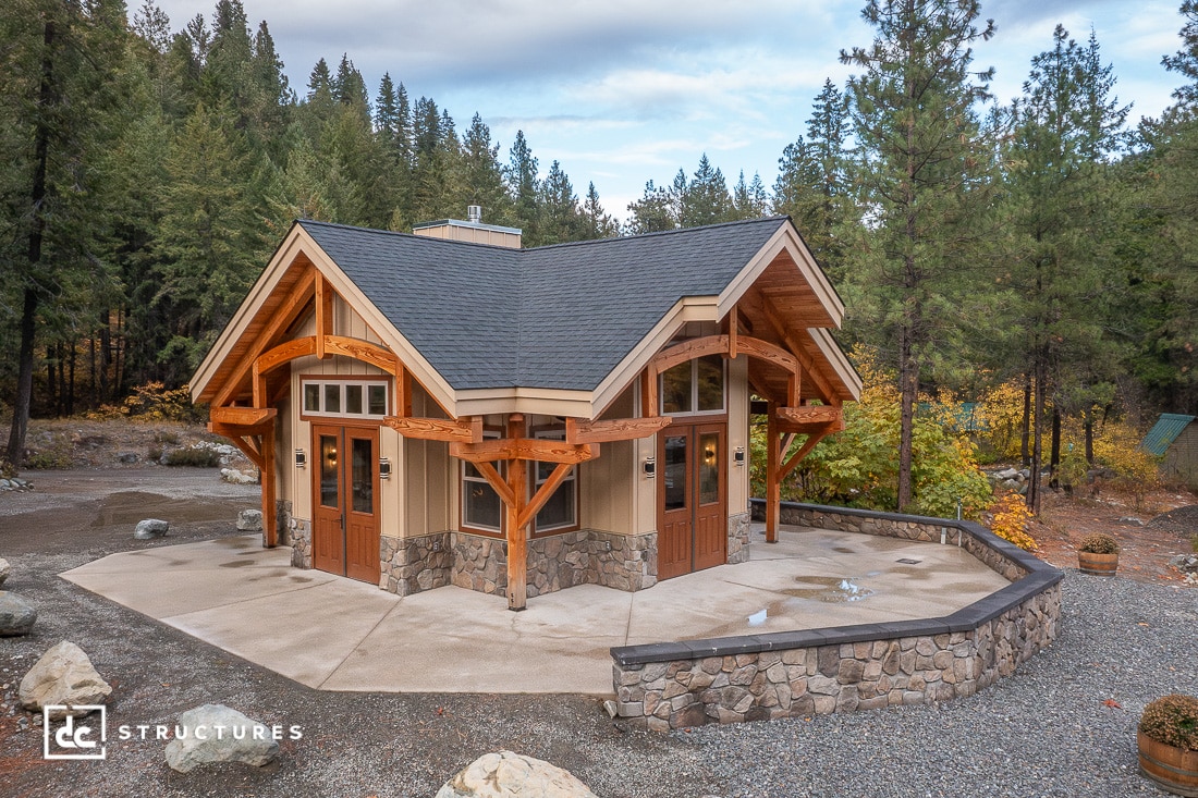 A small modern cabin with wood and stone exterior sits in a forest clearing, surrounded by tall pines and autumn foliage.