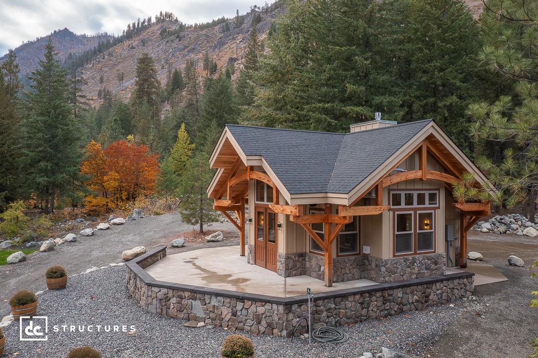 A small modern cabin with timber beams and stone accents sits among pine trees and mountains, with large windows and a curved patio.