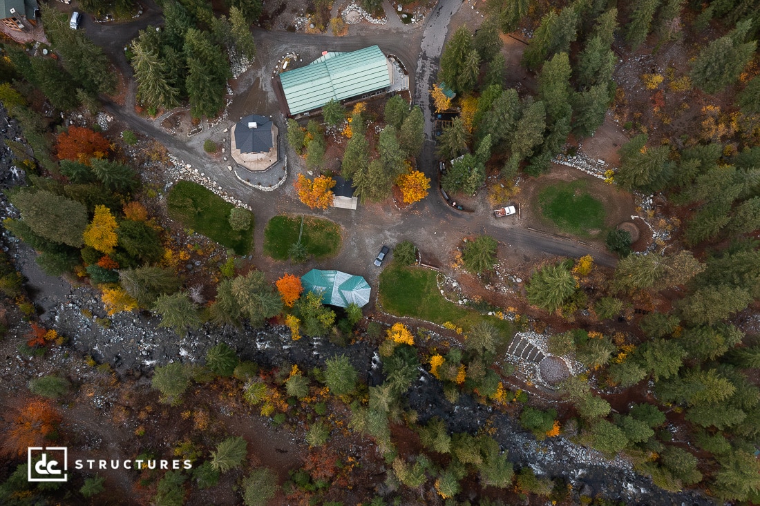 Aerial view of a forested property with multiple buildings, winding roads, scattered vehicles, and colorful autumn trees. A river runs along the bottom edge.