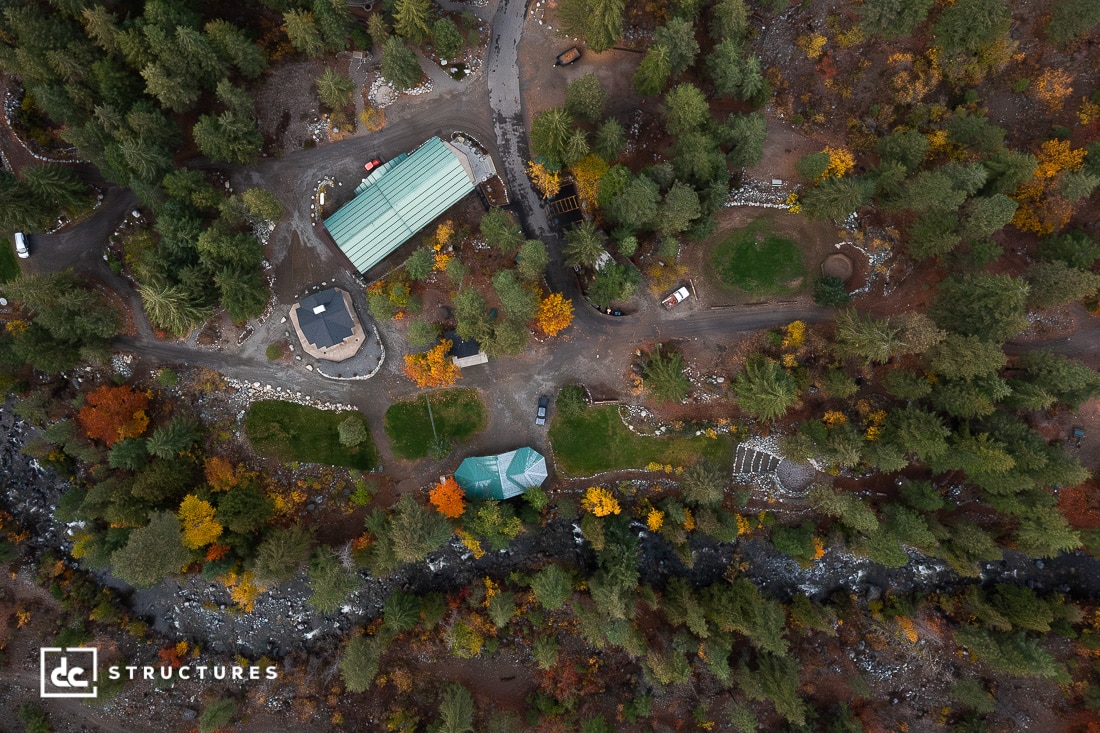 Aerial view of a forested area with buildings, grassy lawns, paths, and a river running through the bottom; trees display autumn colors.