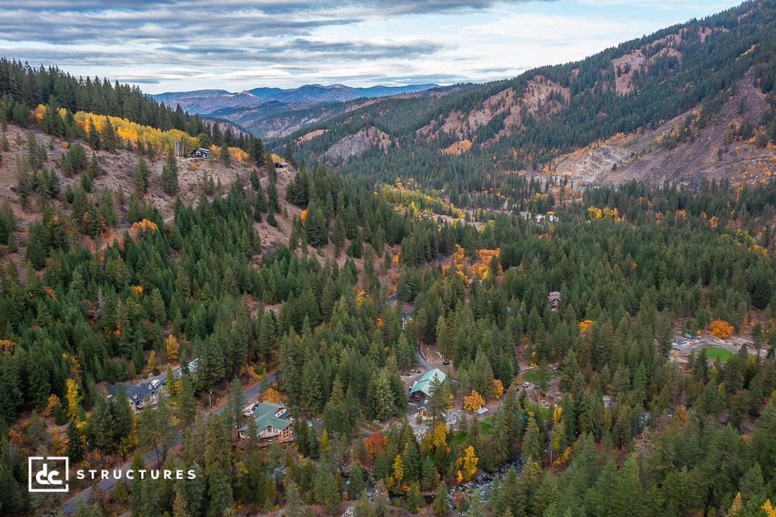 Aerial view of a forested mountain valley with scattered homes among dense evergreen trees, autumn foliage, and rolling hills under a cloudy sky.