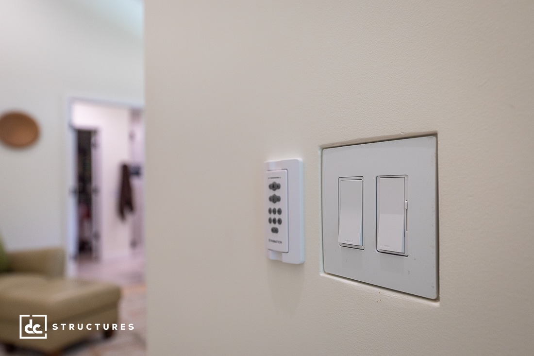Close-up of two light switches and a remote control on a beige wall; in the background, a living space with a green chair and blurred decor.