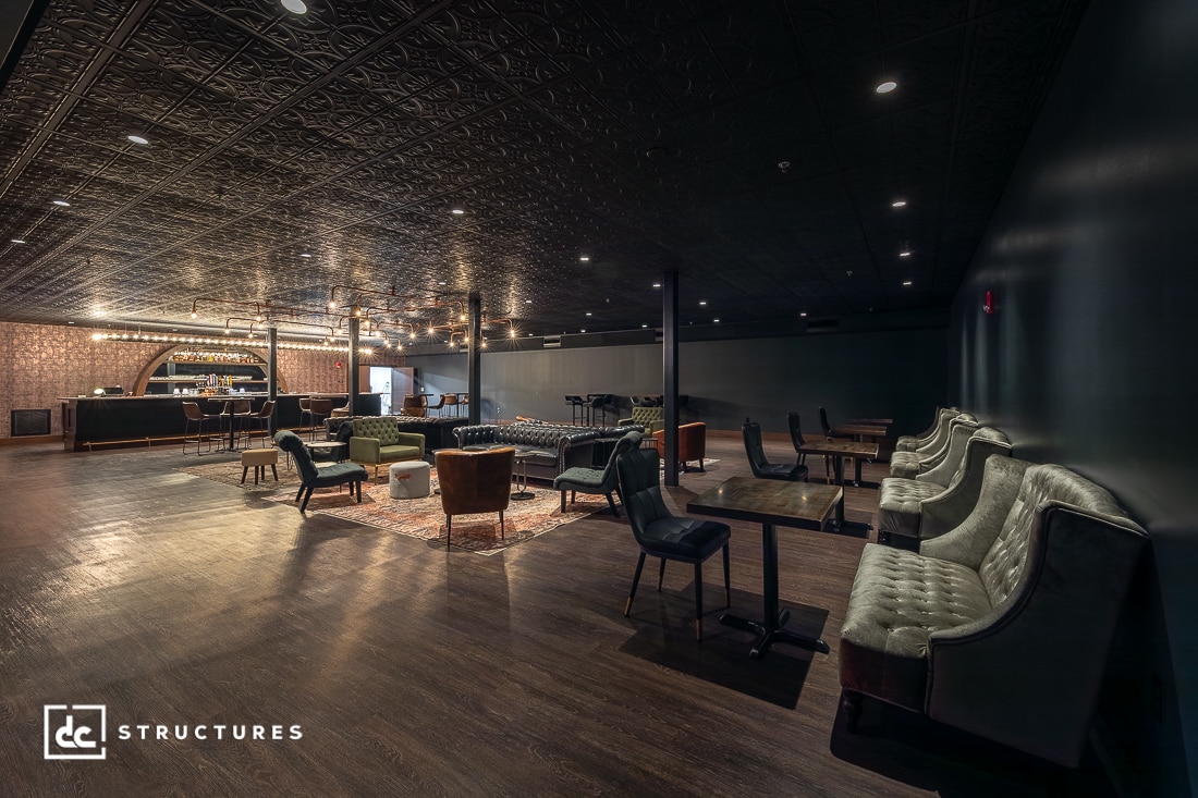 A stylish lounge with dark wood floors, tufted sofas, modern chairs, low tables, and a bar in the back. Warm lighting creates a cozy ambiance. The ceiling features decorative patterns.
