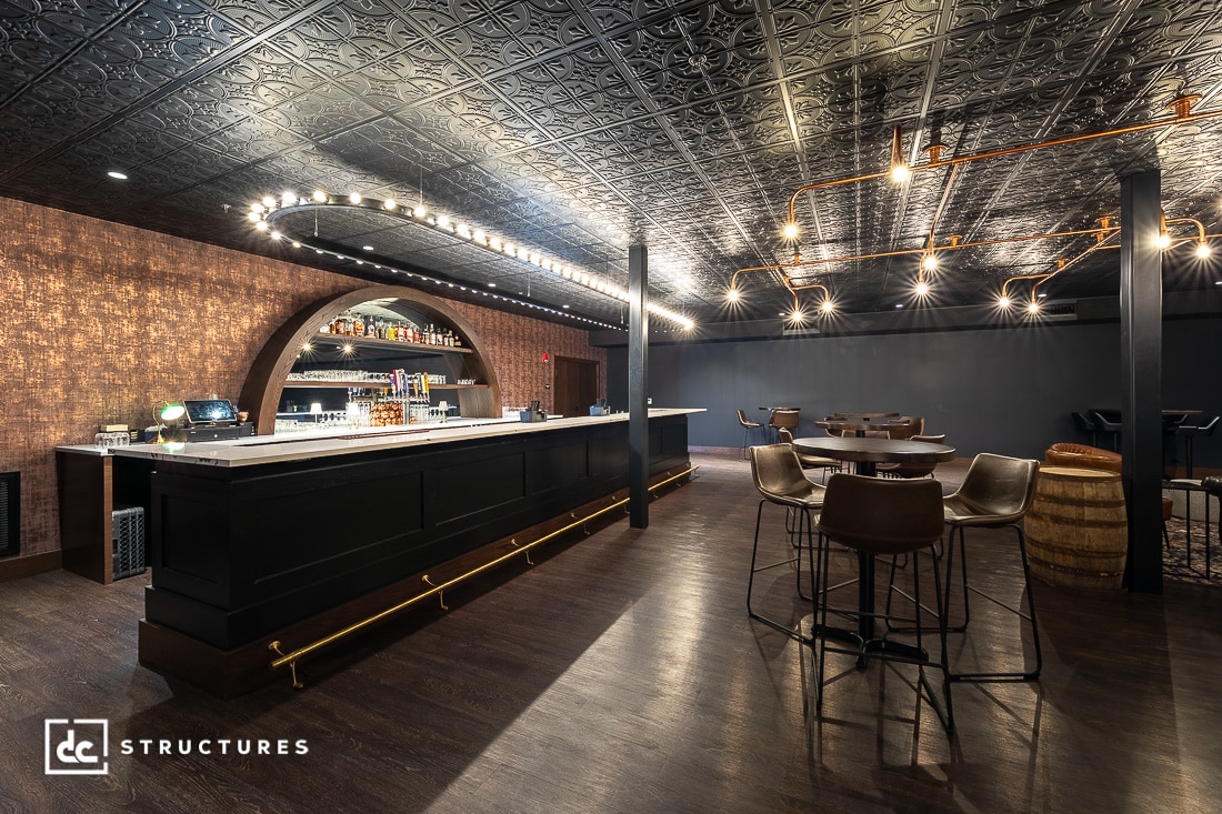 A stylish bar with a black counter, arched liquor shelves, modern lighting, dark wood floors, patterned black ceiling, and round tables.