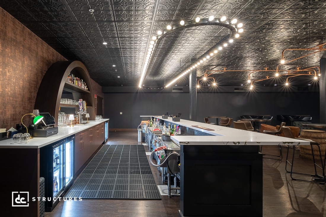 A modern bar with a curved counter, backlit shelves, and rows of drinks. Ornate black ceiling tiles, dramatic lighting, lounge chairs, and tables in the background.