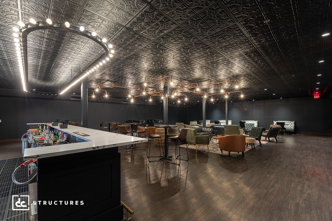 A modern lounge with a dark ornate ceiling, circular chandeliers, mixed seating, a sleek bar in front, and polished wooden floors.
