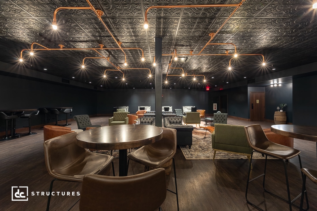 A modern lounge with leather chairs, round wooden tables, patterned rugs, exposed copper ceiling pipes, and warm pendant lights.