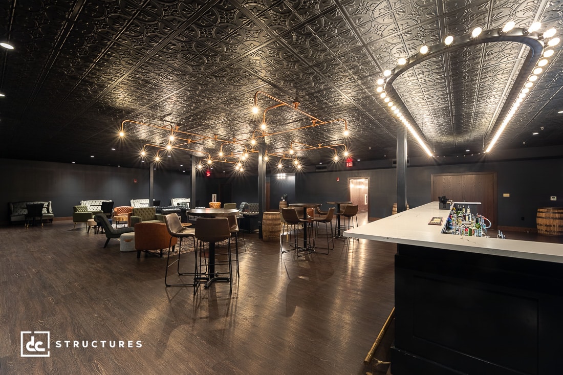 A spacious, stylish lounge with dark walls and an ornate ceiling, modern lighting, a white bar, assorted seating, barrel tables, and polished wood floors. The atmosphere is cozy and sophisticated.