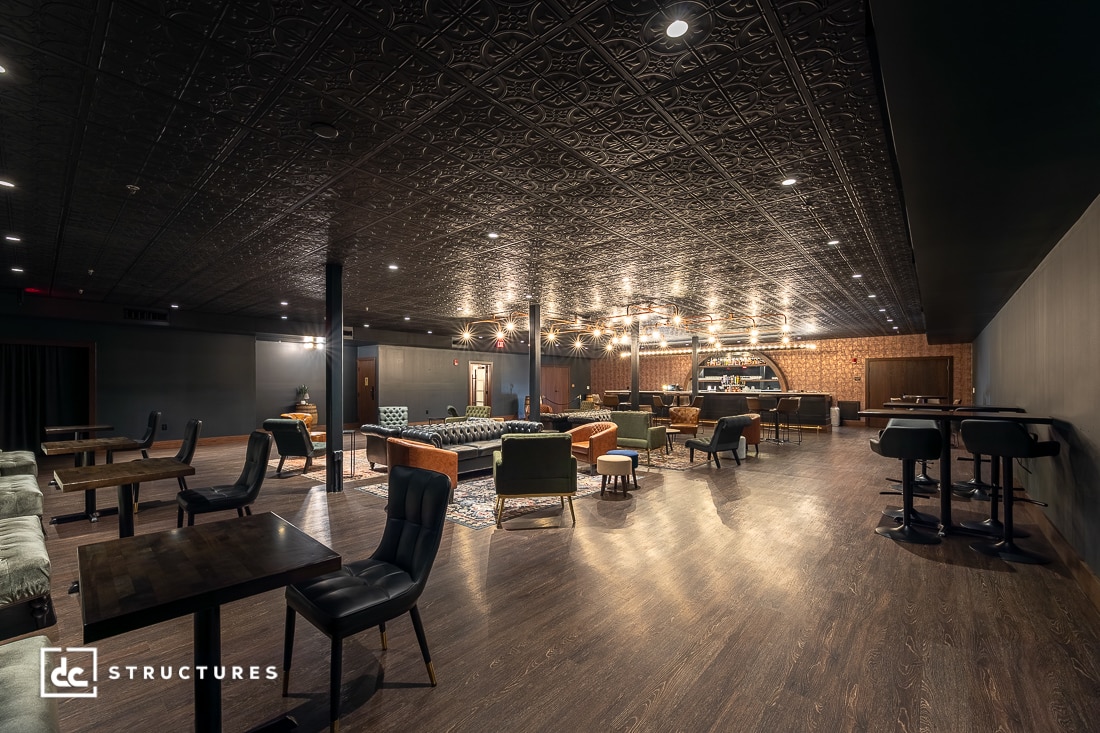 Spacious, modern lounge with dark wood floors, patterned ceiling, mixed seating including sofas and chairs, round bar area in the back, and warm lighting.