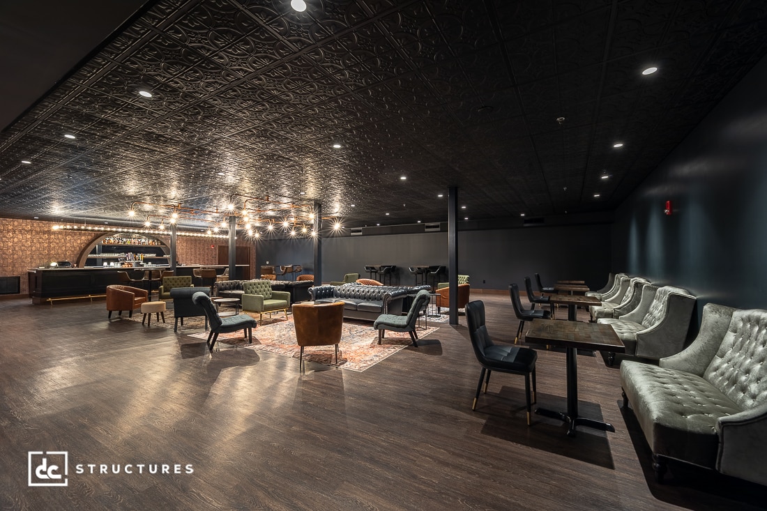 A stylish lounge with vintage armchairs, sofas, and rugs, a dark patterned ceiling, wooden floors, bar seating, and warm lighting.