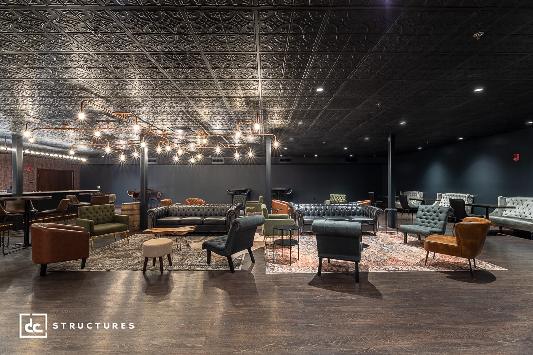 Modern lounge with eclectic seating, including armchairs, sofas, and small tables on patterned rugs. Decorative ceiling, dark walls, and artistic chandeliers create a cozy, upscale atmosphere.