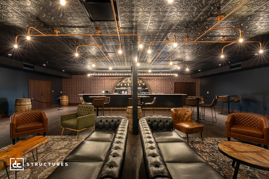 A stylish lounge with black leather sofas, armchairs, and a patterned ceiling features warm custom lighting and a barrel table.